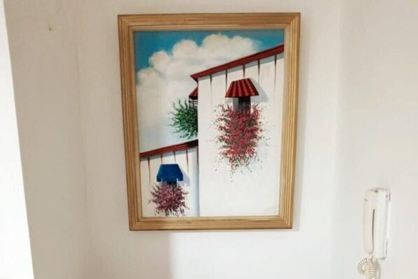 A framed artwork of a building with a red roof and a blue awning, hanging on a plain wall.
