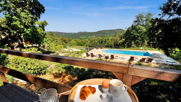 A wooden deck with a table set for breakfast, overlooking a swimming pool and lush greenery.