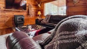 L’intérieur chaleureux d’un chalet avec un plafond en bois, une tête de cerf accrochée au mur, une cheminée, un canapé en cuir et un téléviseur.