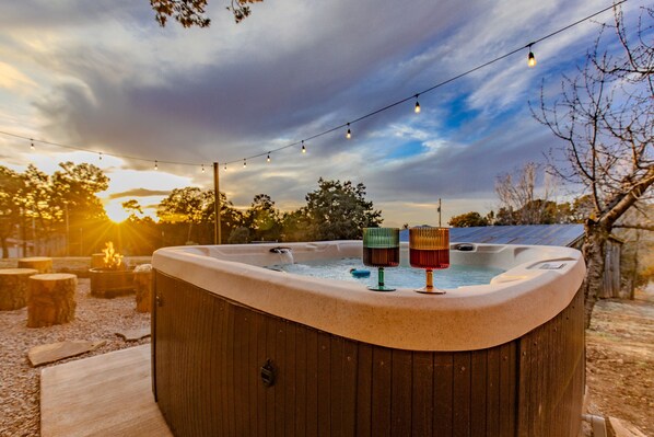 Un jacuzzi con dos vasos coloridos, rodeado de taburetes de madera y luces de cuerda, con árboles y un cielo nublado de fondo.
