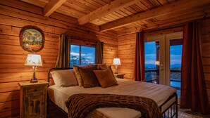 Une chambre confortable, aménagée dans un chalet en bois, avec un grand lit, deux lampes de chevet, un tableau encadré au mur et une vue sur les montagnes par la fenêtre.
