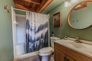 Baño con cortina de ducha que muestra un paisaje boscoso, inodoro, lavamanos con espejo y armarios de madera.