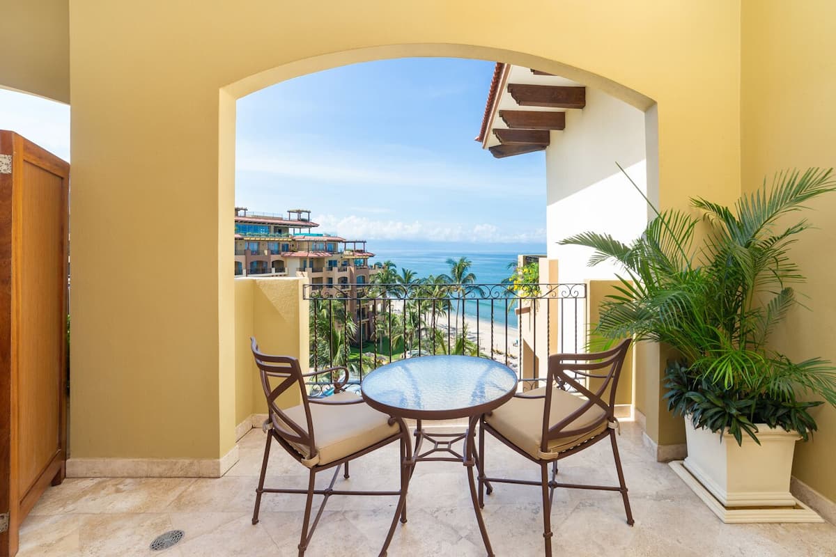 Un balcon avec une table ronde et des chaises, donnant sur une plage et un ensemble d’immeubles.