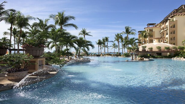 A large swimming pool with palm trees and a multi-story hotel building in the background.