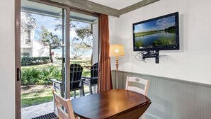 Un área de comedor con una mesa y sillas, un televisor montado en la pared, y vistas de un jardín a través de un ventanal.