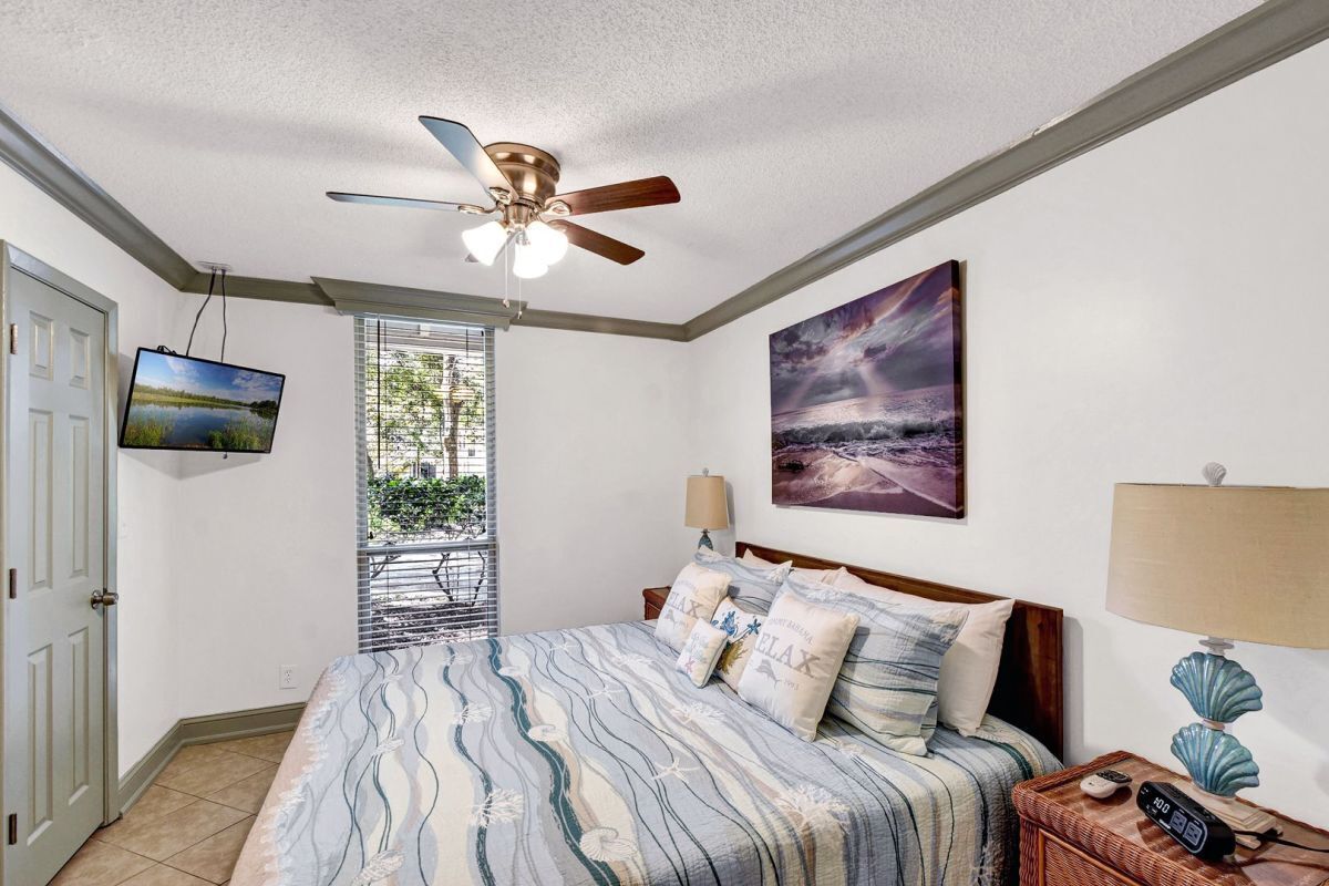 A bedroom with a bed, a ceiling fan, a TV, a painting, a window, and a nightstand.