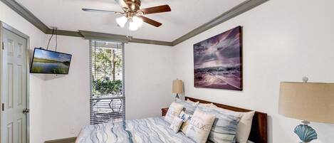 A bedroom with a bed, a ceiling fan, a TV, a painting, a window, and a nightstand.