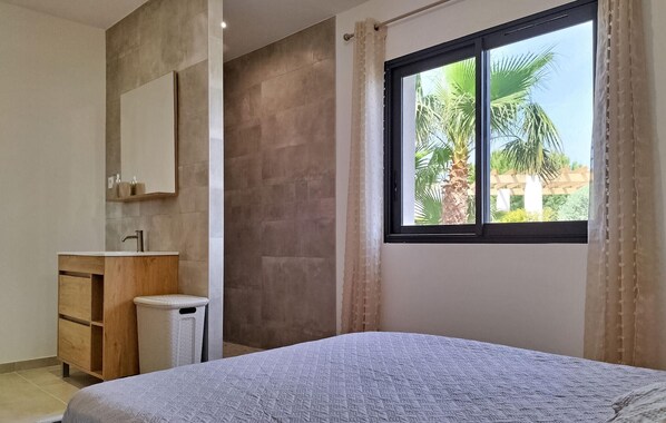 A bedroom with a bed, a wooden cabinet, a mirror, and a window with a view of palm trees.