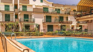 Une piscine entourée d’une terrasse carrelée, elle-même bordée d’un immeuble à plusieurs étages doté de balcons et de volets verts.
