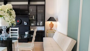 Un salon moderne avec une table à manger en verre, des chaises blanches, un canapé beige, une petite table d’appoint avec une lampe et un meuble noir avec des étagères.