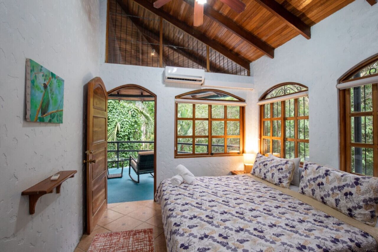 Une chambre dotée d’un grand lit, de poutres apparentes en bois au plafond et de nombreuses fenêtres donnant sur un paysage verdoyant.