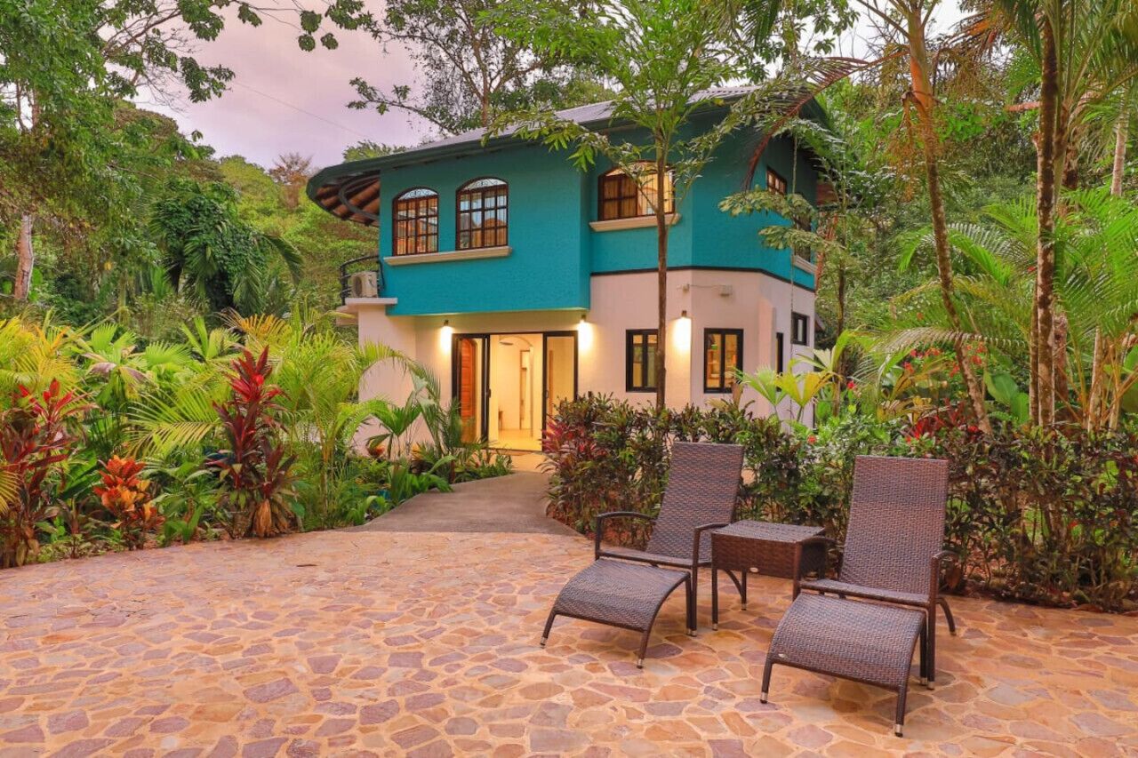 Une maison à deux étages dotée d’une terrasse en pierre, entourée d’une végétation luxuriante et d’une entrée bien éclairée.