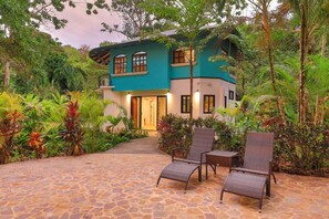 Een huis met twee verdiepingen, een stenen terras, omringd door weelderige begroeiing en een goed verlichte entree.
