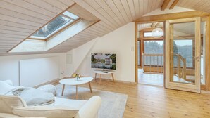 Quarto de loft aconchegante com teto inclinado, vigas de madeira, claraboia, janela grande, TV, mesa pequena com um vaso de flores e varanda com vista para o exterior.