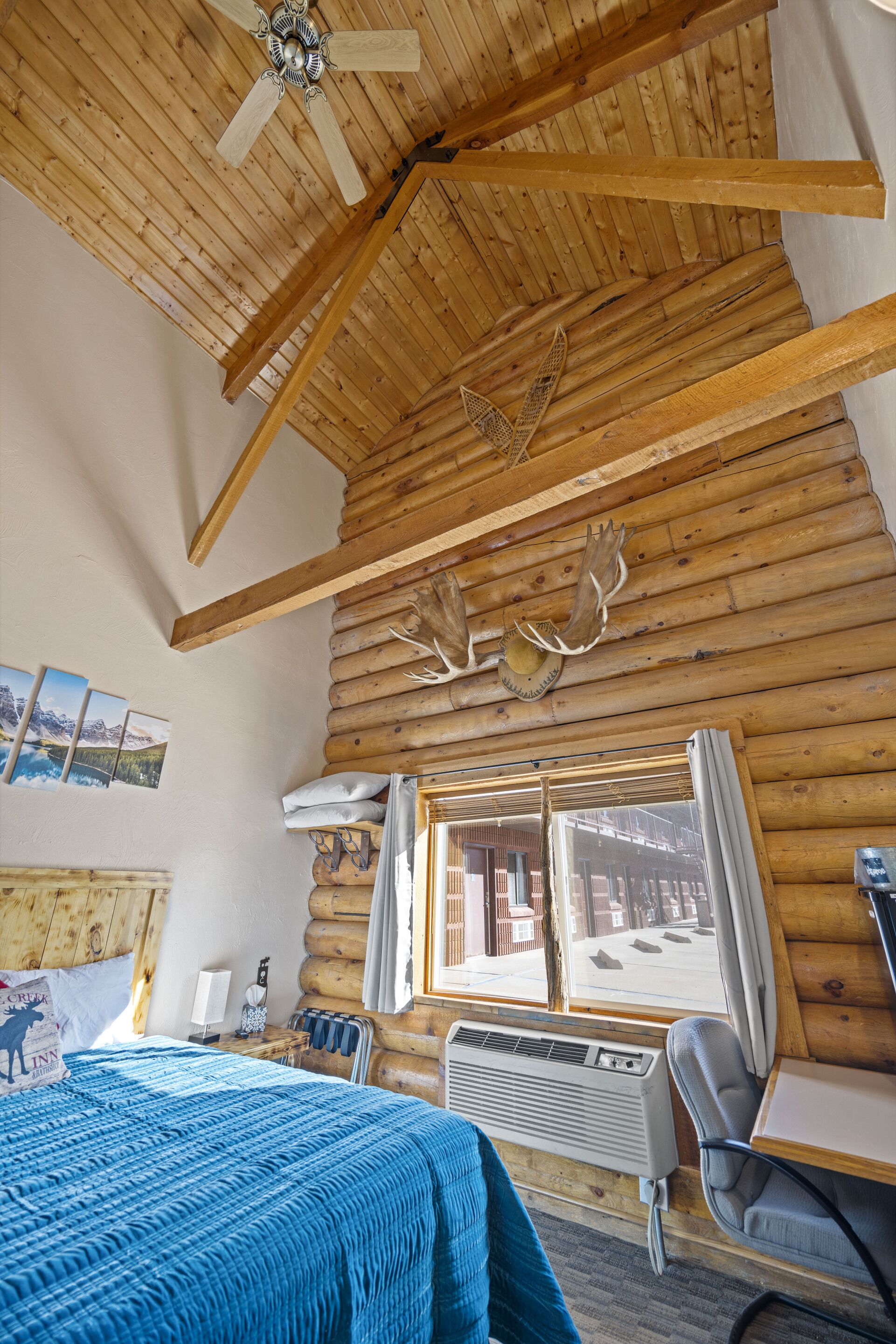 A log cabin room with a bed, a desk, a chair, a window with curtains, a ceiling fan, and a mounted deer head.