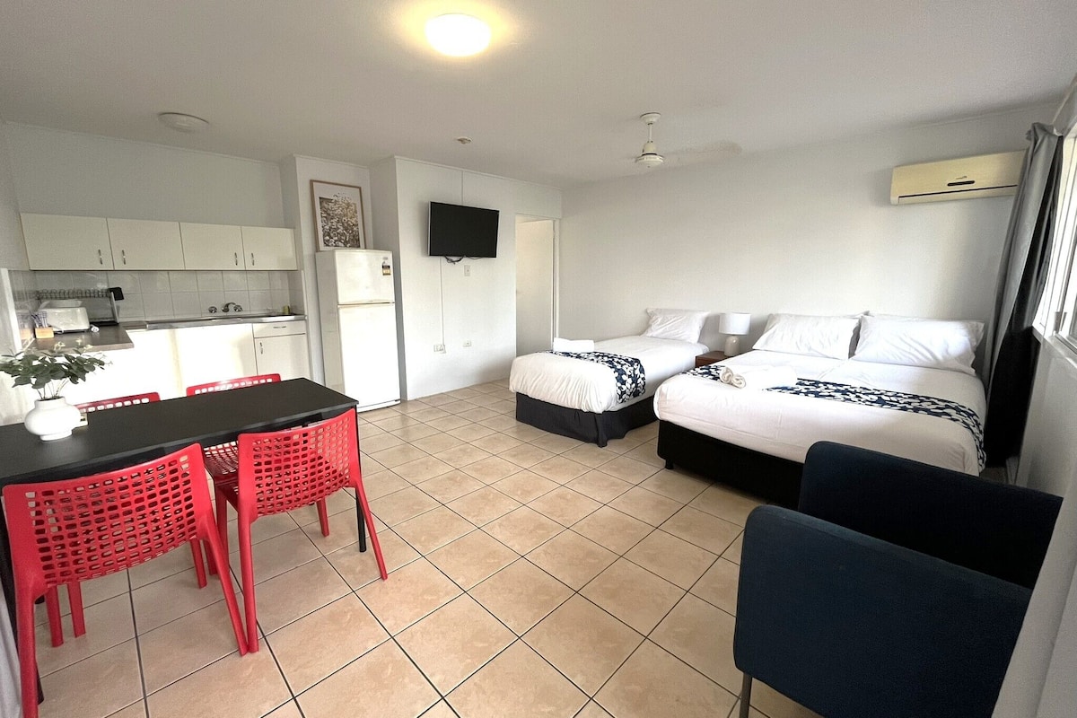 Una habitación de hotel compacta con dos camas, una kitchenette, una zona de comedor con sillas rojas y un televisor montado en la pared.