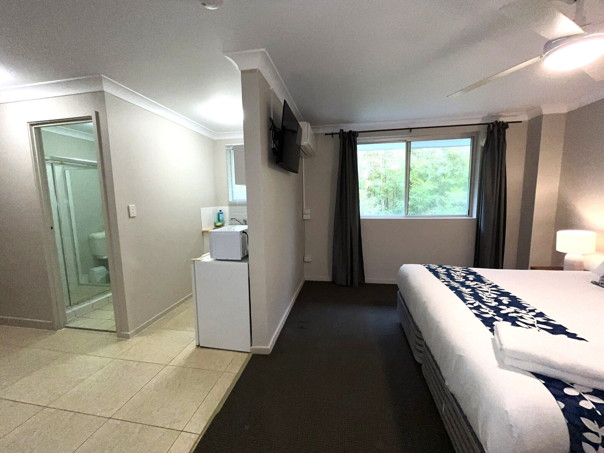 A hotel room with a bed, a small desk, a window with curtains, and a bathroom visible through an open door.