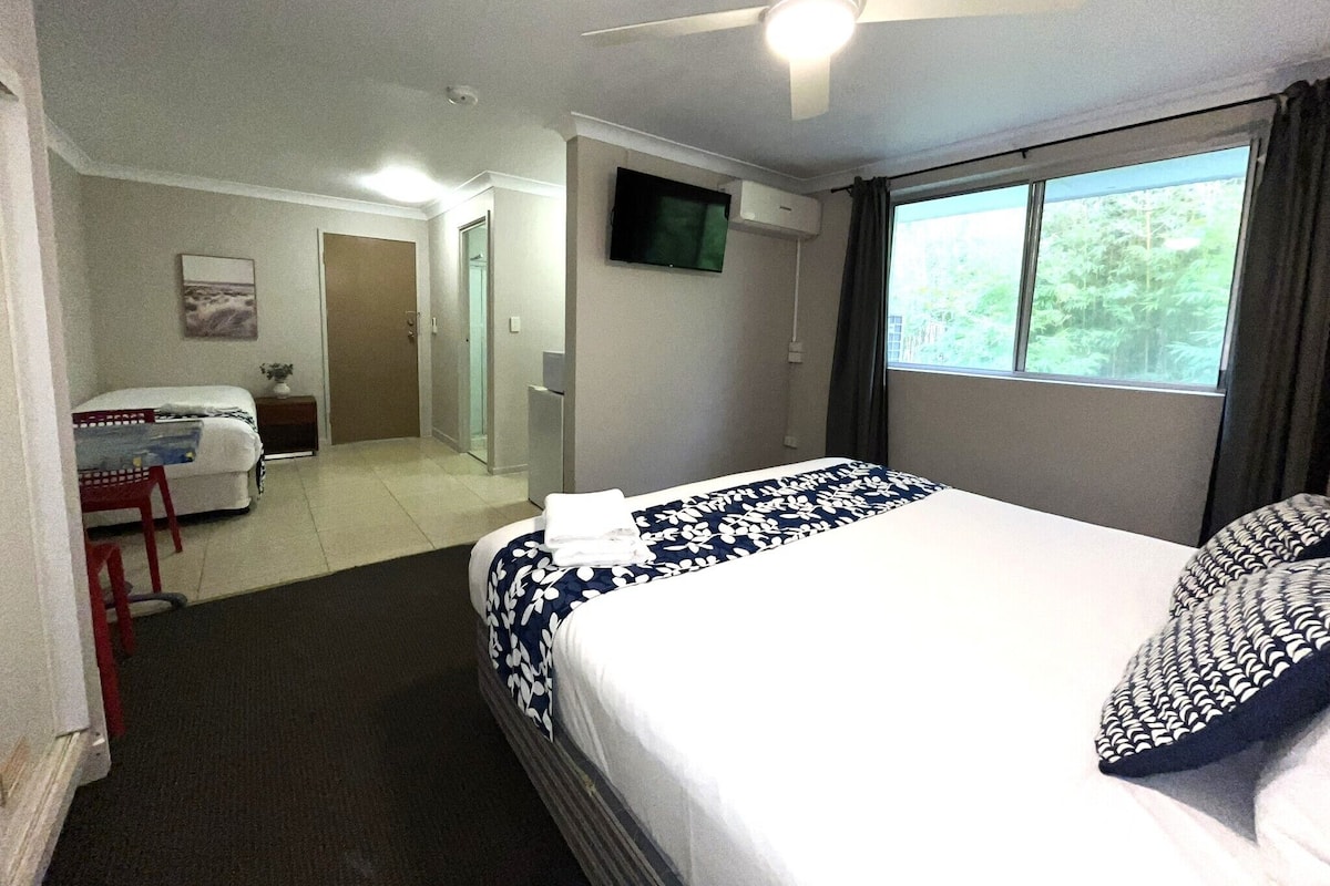 Habitación de hotel con cama, escritorio, silla, televisor y ventana con vista a follaje.