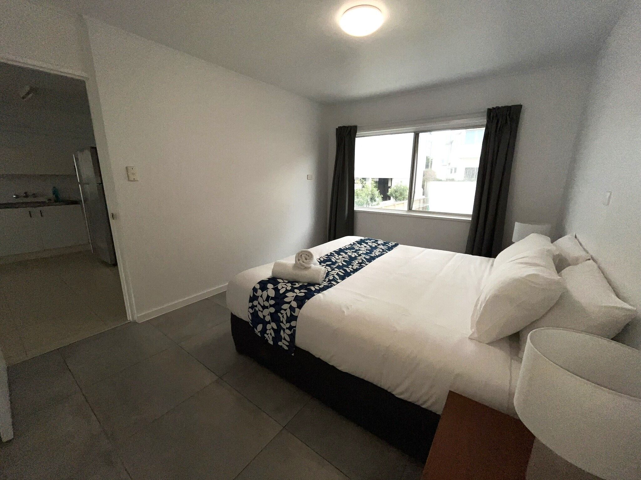 Hotellihuone, jossa on sänky, ikkuna verhoilla, pieni pöytä ja pieni keittiöalue.