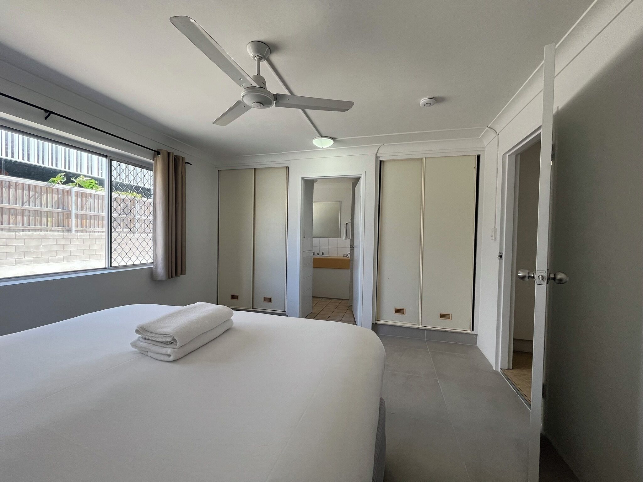 Une chambre d’hôtel avec un lit, un ventilateur de plafond, une salle de bain avec un lavabo et une vue sur l’extérieur par une fenêtre.