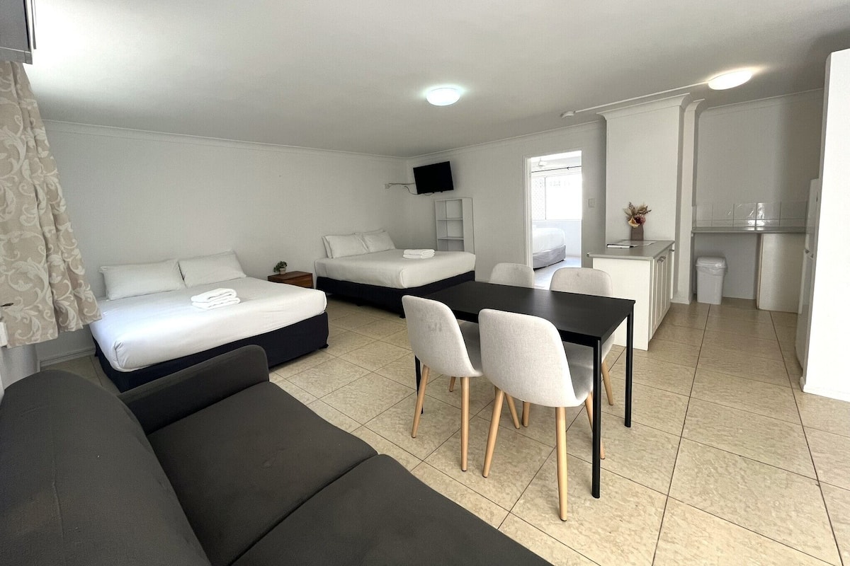 Una habitación moderna y bien iluminada con un sofá, dos camas, una mesa de comedor con sillas, una pequeña cocina y un televisor montado en la pared.