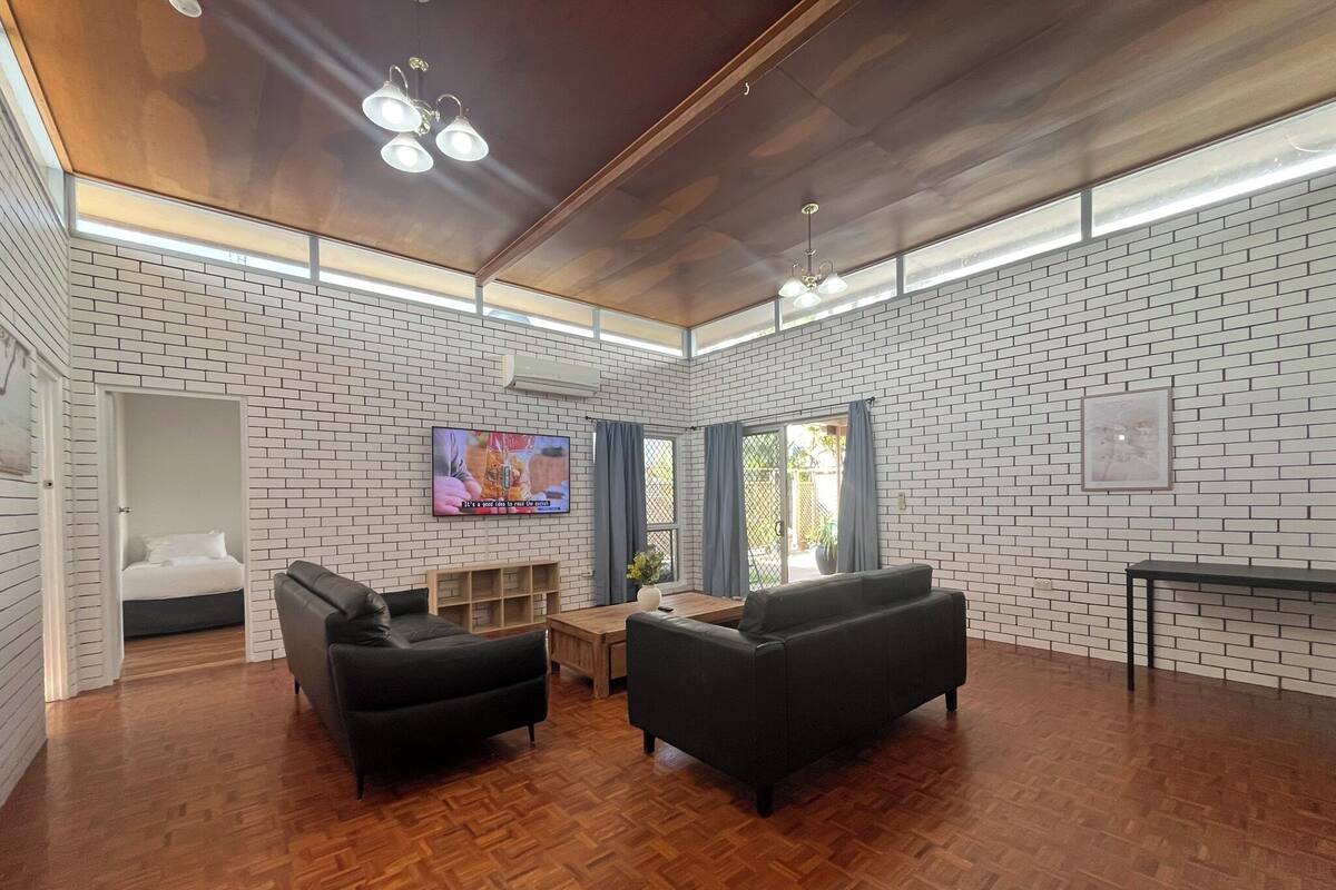 Un salón amplio con suelo de madera, un televisor montado en la pared y una cama visible a través de un umbral abierto.
