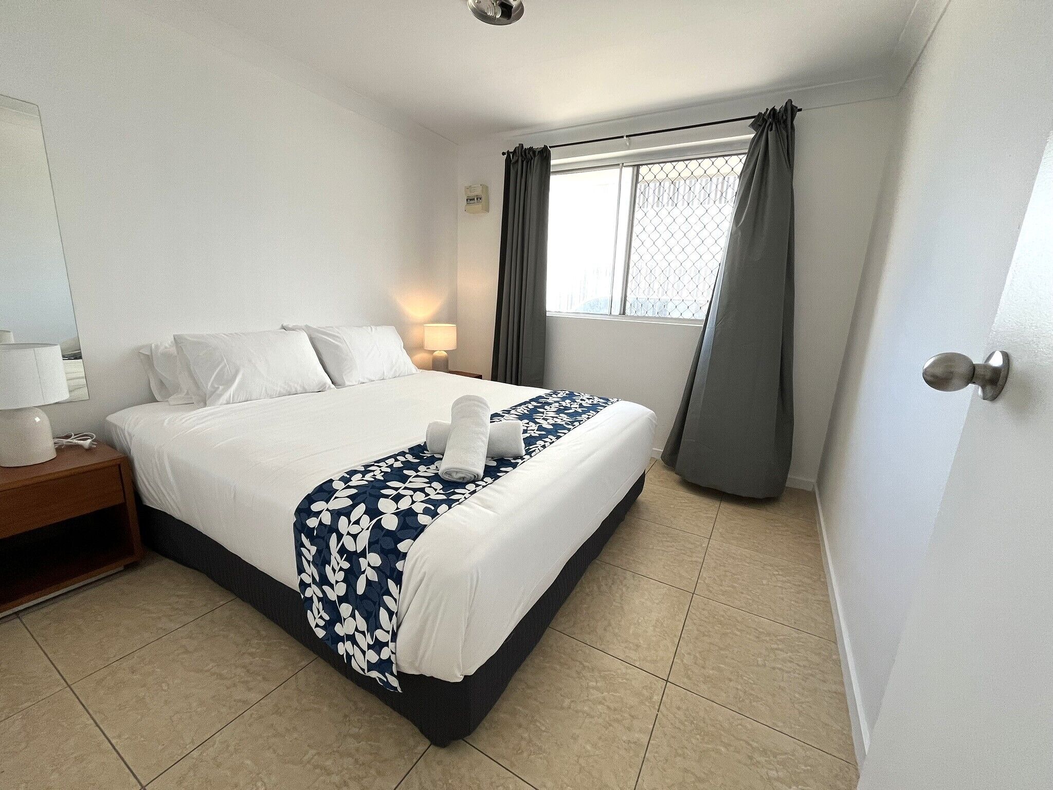 Une chambre d’hôtel avec un lit bien fait, une lampe de chevet, une fenêtre avec des rideaux et une porte dotée d’une poignée.