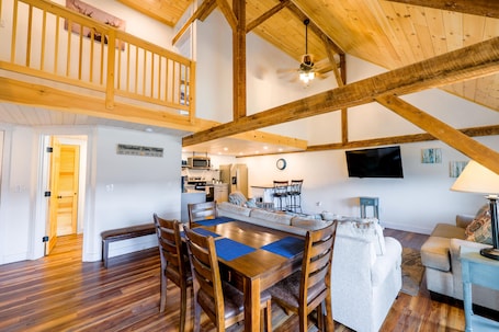 A spacious living area with a wooden dining table, high ceilings with exposed beams, a kitchen area, and a balcony.