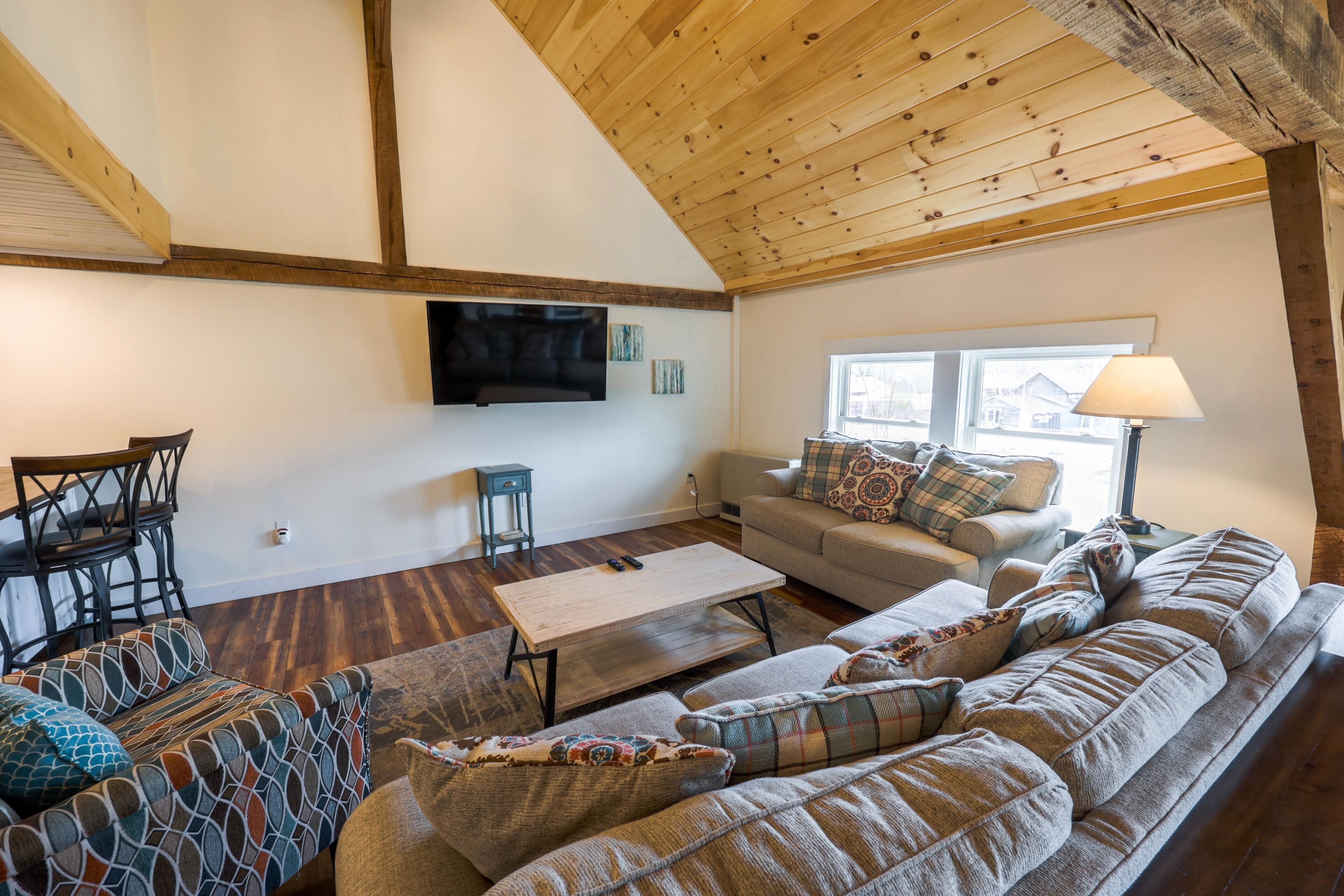Un accogliente soggiorno con soffitto in legno, una TV a schermo piatto montata a parete, un divano, un tavolino da caffè e una finestra con una lampada.