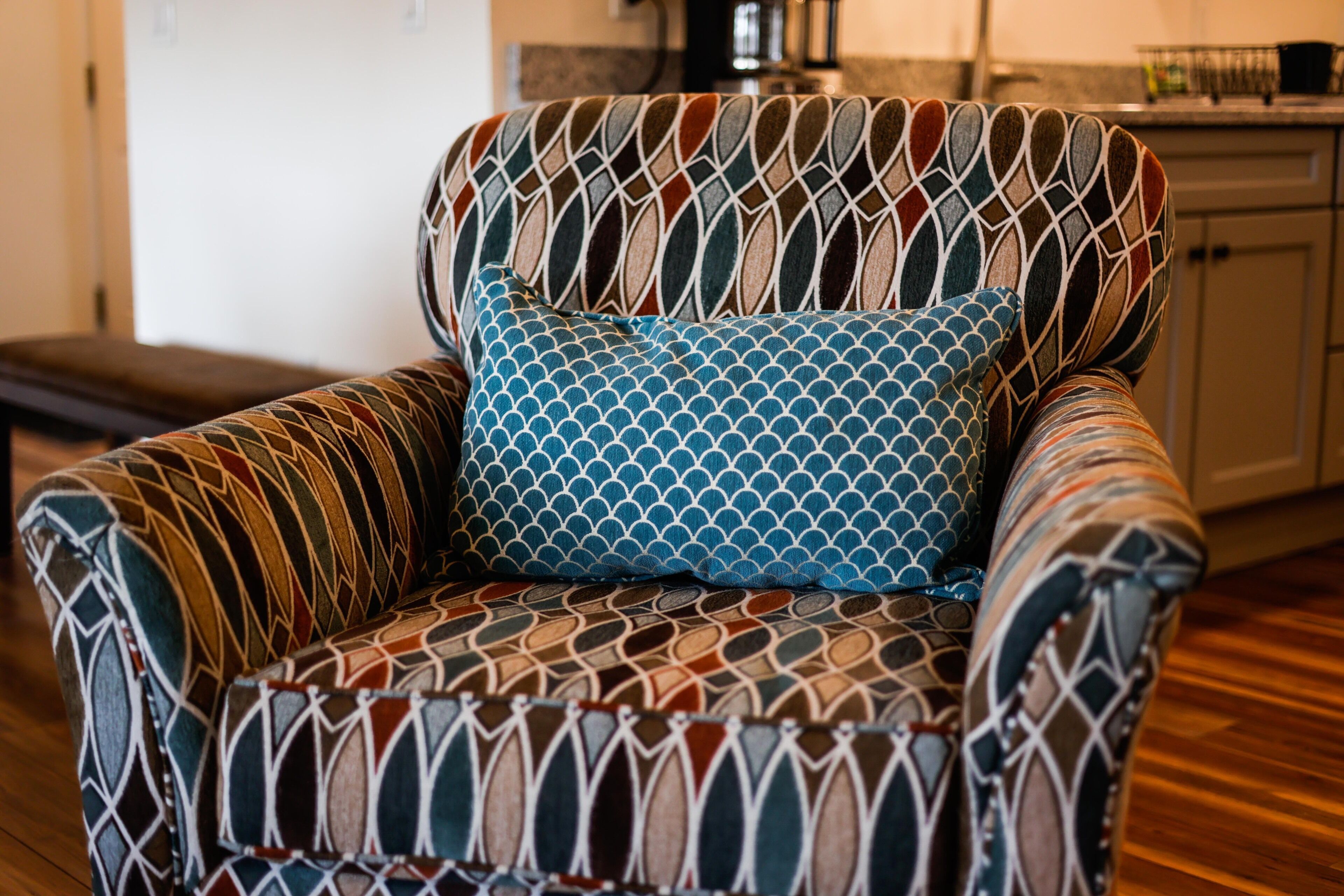 A patterned armchair with a matching cushion in a room with wooden flooring and a kitchen in the background.