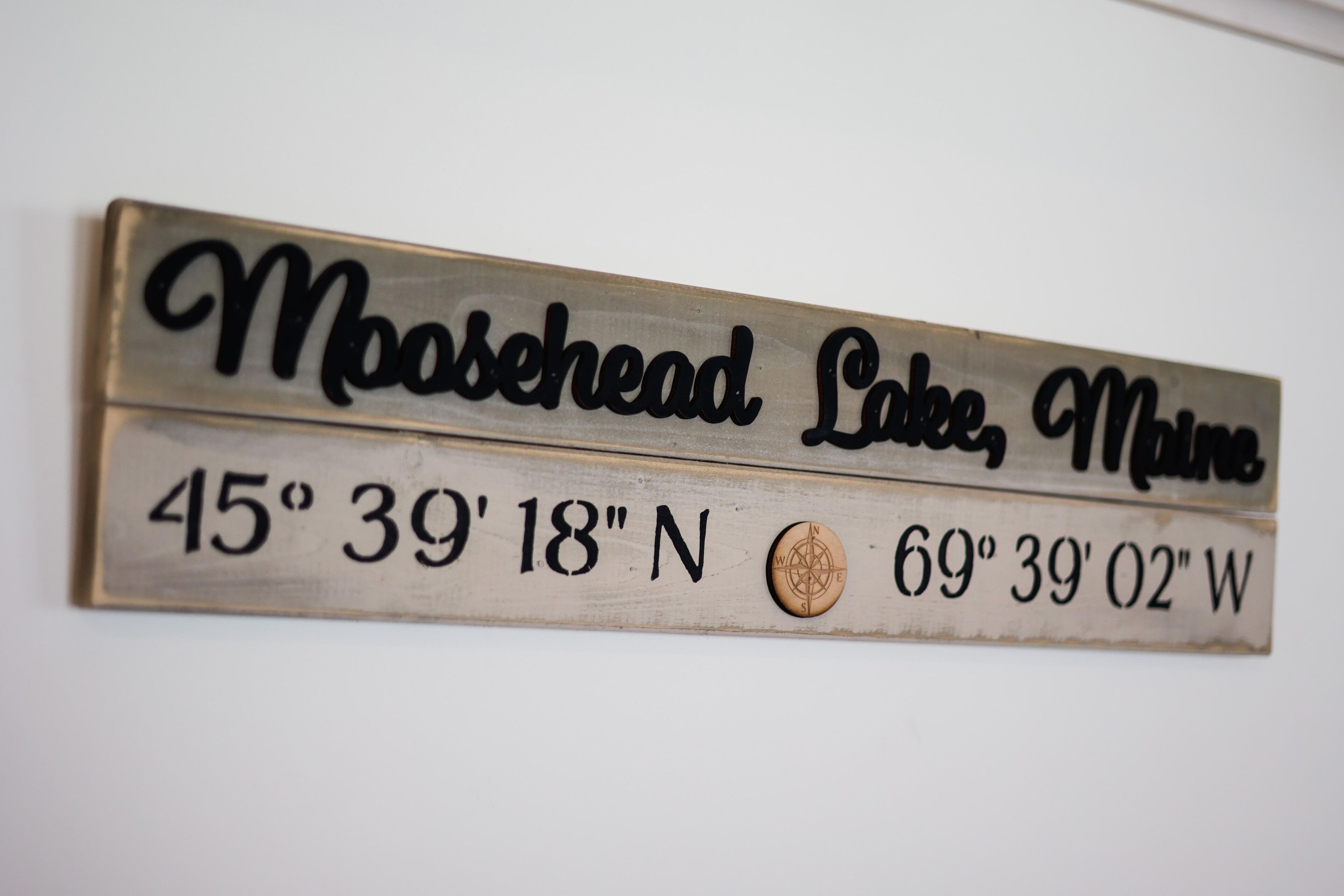 A metal sign with the text "Moosehead Lake, Maine" and coordinates "45° 39' 18" N, 69° 39' 02" W".