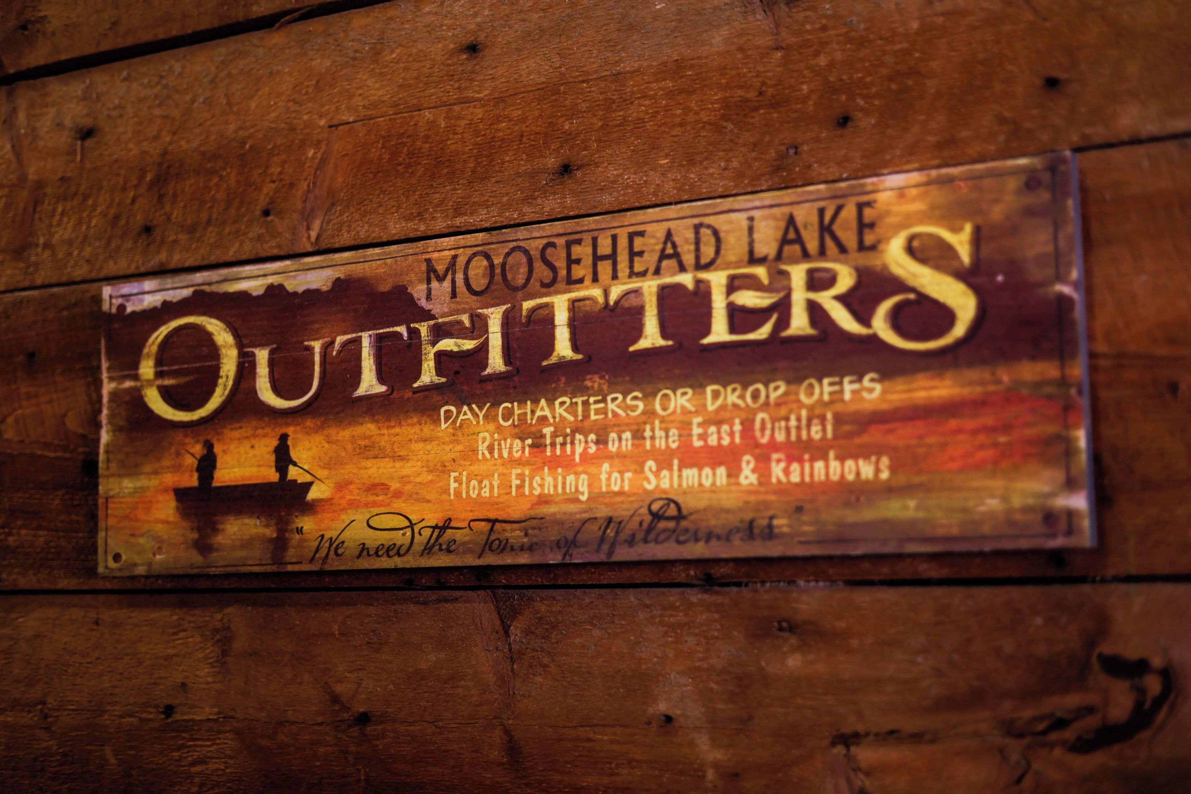 Une pancarte en bois portant l’inscription « Moosehead Lake Outfitters » et mentionnant diverses activités, telles que les excursions d’une journée, les départs en bateau, les excursions en rivière et la pêche à la mouche pour la pêche au saumon et à la truite arc-en-ciel.