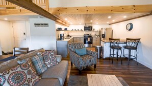 A spacious living area with a sofa, armchair, and a dining table with bar stools. A kitchen with modern appliances is visible in the background. - Historic Barn Suites (Greenville)