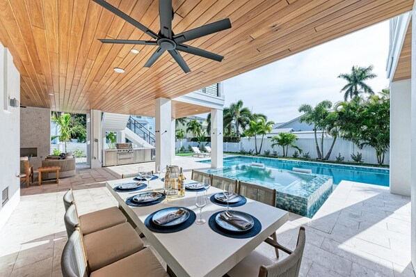 Uma área moderna de refeições ao ar livre com piscina, teto de madeira e vista para a piscina e vegetação.