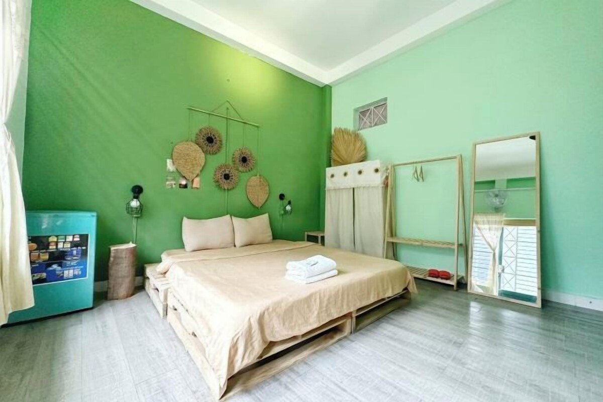 Una habitación con pared verde, cama con ropa de cama color beige, una mesita con espejo y un armario azul.