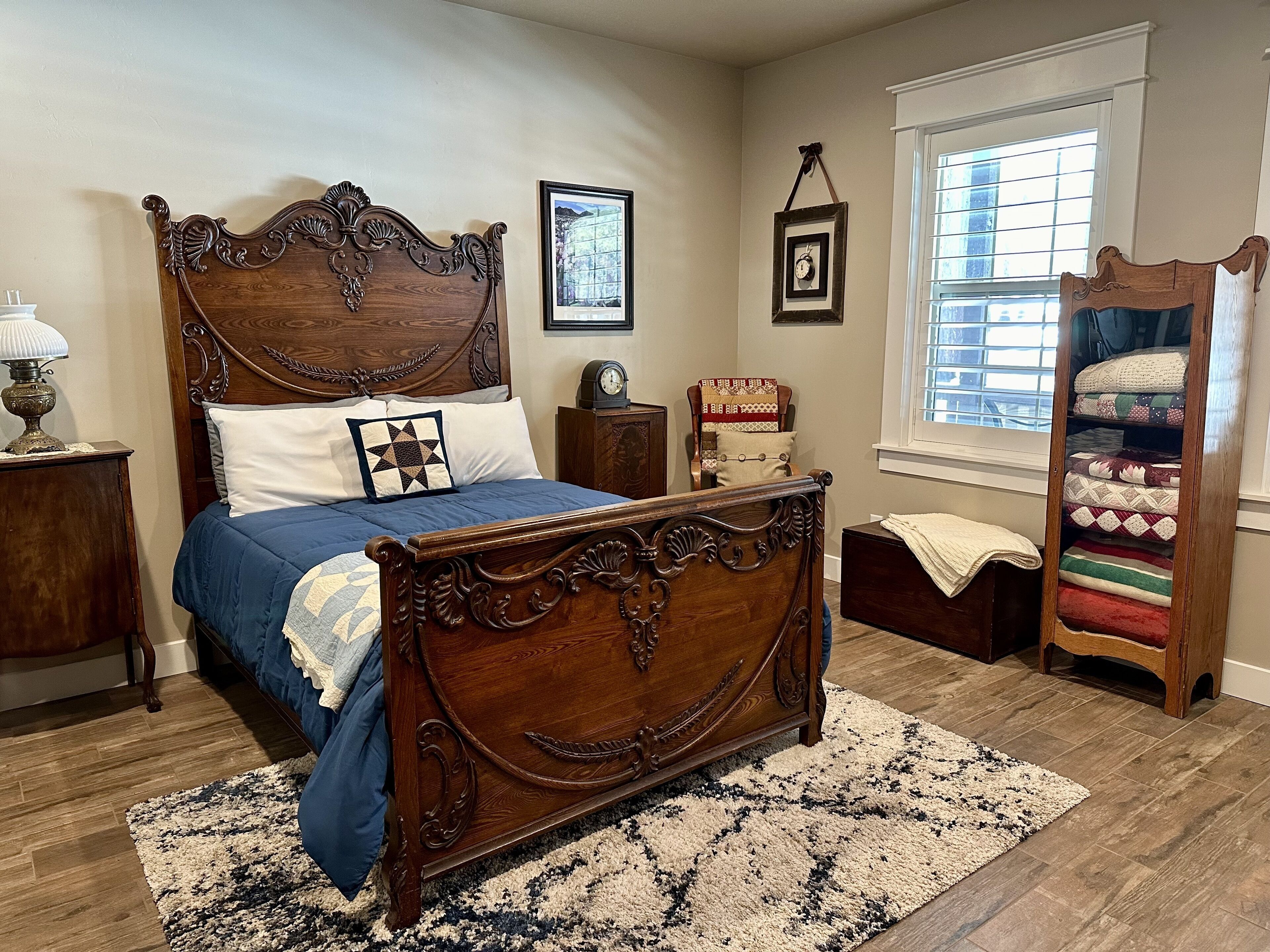 A bedroom with a large wooden bed, a nightstand, a dresser, a window with blinds, and a quilted shelf unit.