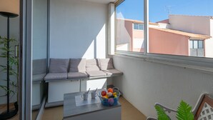 Een balkon met een zachte zithoek, een klein tafeltje met een fruitmand, en uitzicht op omliggende gebouwen.