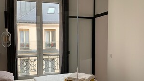 Un dormitorio moderno con una cama grande, un separador de vidrio y vistas a un edificio con balcones.
