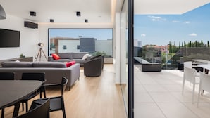 A modern living room with a large window, a sofa, a dining table, and a balcony with outdoor furniture. - Old Olive IV by Interhome (Pula/Vodnjan)