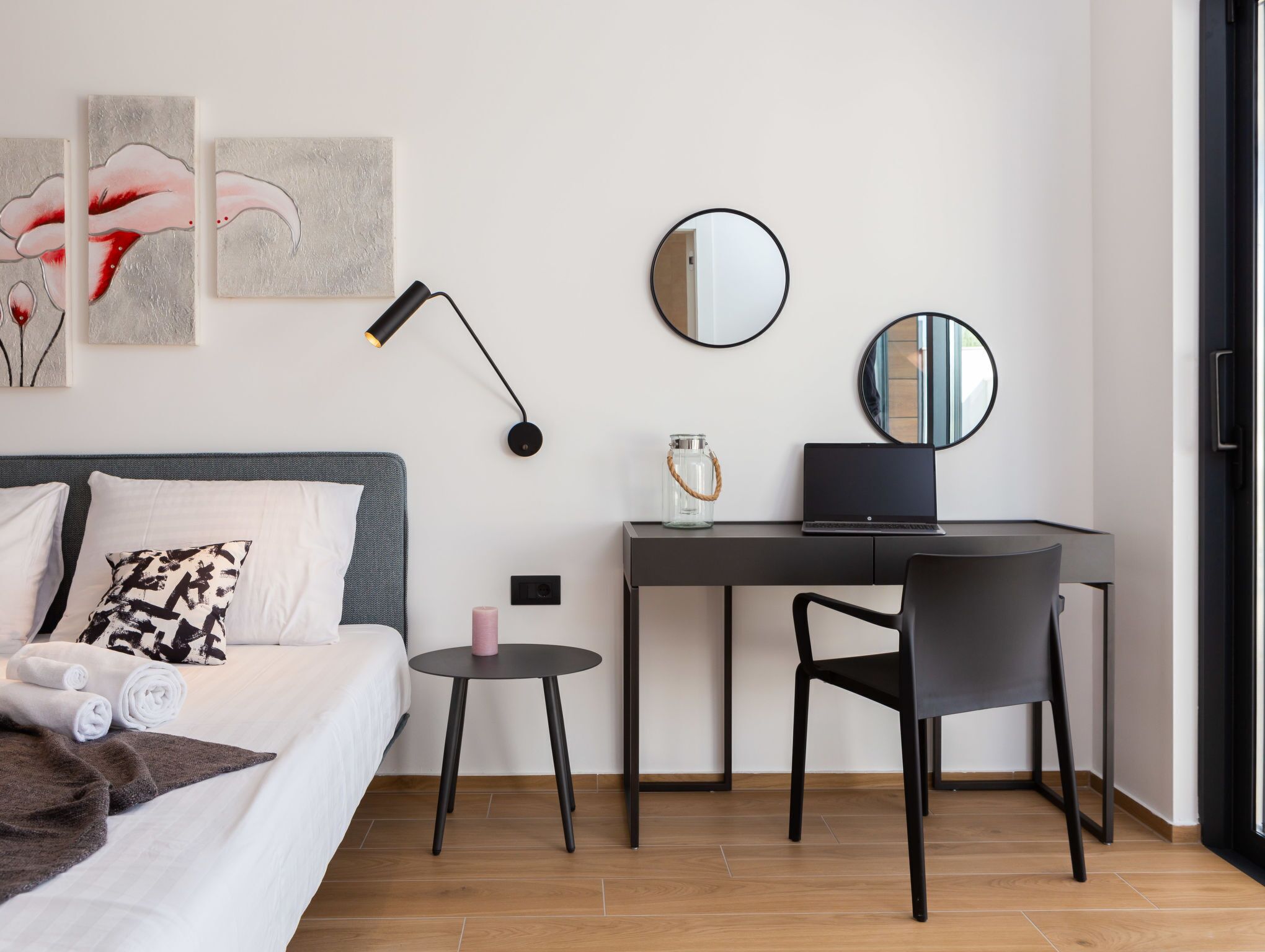A modern hotel room with a bed, a desk, a chair, a round mirror, and artwork on the wall.