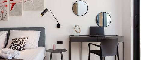 A modern hotel room with a bed, a desk, a chair, a round mirror, and artwork on the wall.