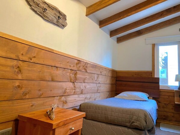 A bedroom with a wooden headboard, a bed with a quilted bedspread, a nightstand with a lamp, and a window with daylight.