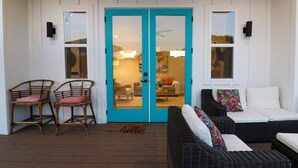 Una terraza de madera con mobiliario de mimbre y una puerta azul que da a un espacio interior.