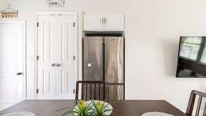 Comedor con una mesa, sillas, un florero con flores, un televisor montado en la pared y un refrigerador de acero inoxidable.