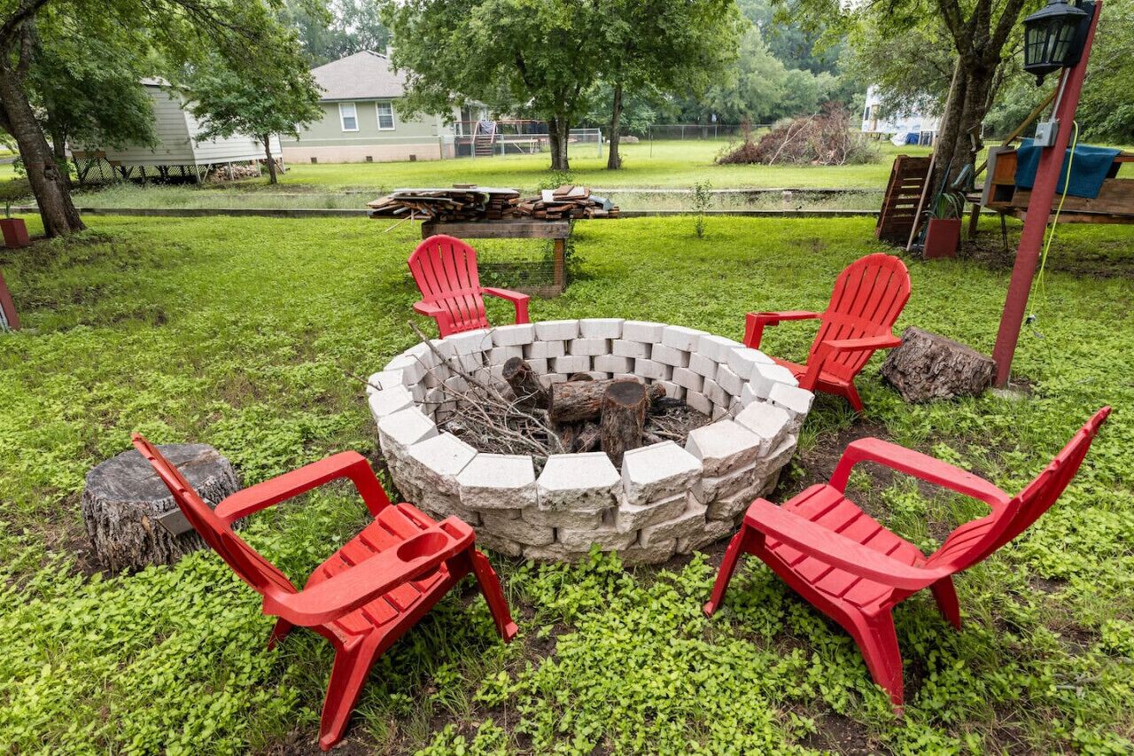 A stone fire pit with four red Adirondack chairs arranged around it in a grassy outdoor area.