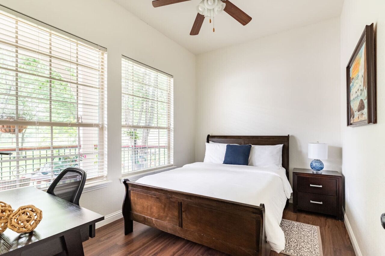 A bedroom with a wooden bed, two windows with blinds, a ceiling fan, a nightstand with a lamp, and a framed picture on the wall.