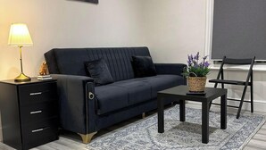 A modern living room with a dark sofa, a side table with a lamp, a small decorative item, a potted plant, and a chair. - Studio - Sleeps 3 - Near Ealing Broadway Station (London)