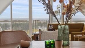 A spacious room with a large window offering a view of the sea and hills. There is a cozy seating area with a sofa and a table with a vase of dried flowers.