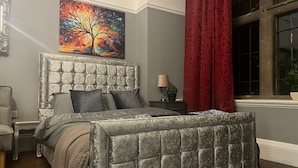 A bedroom with a tufted headboard, a bed with grey bedding, a nightstand with a lamp, a red curtain, and a window with a view. - Immaculate Studio in Bristol (Bristol)