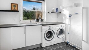 A modern laundry room with a washing machine and dryer, a window with a view of greenery, and a shelf with toiletries. - "Elsi" - 1.3km from the sea by Interhome (Blåvand)